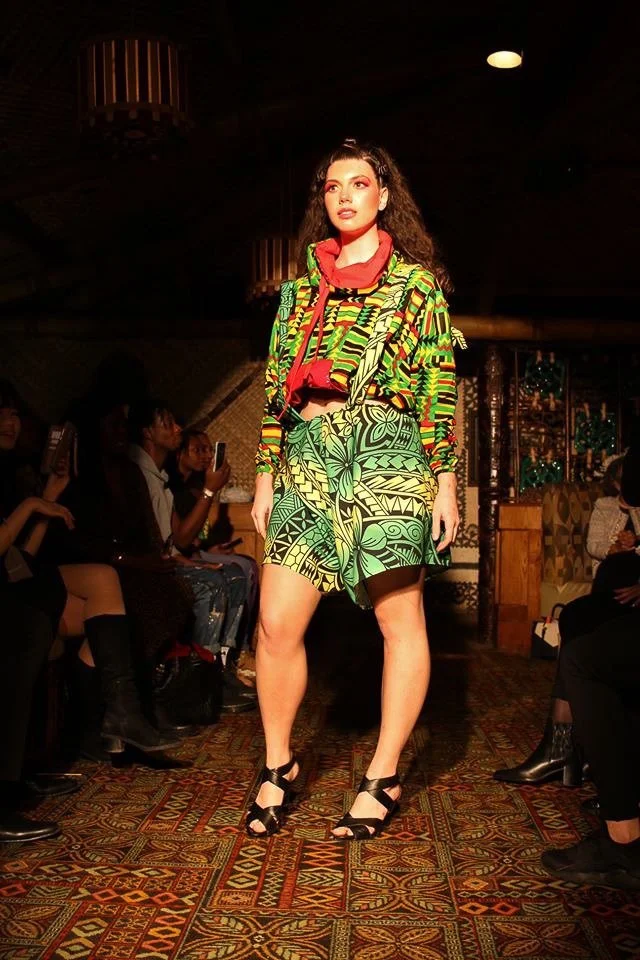 A model poses on the runway wearing a green and yellow patterned skirt with matching top. A red color frames her neck.