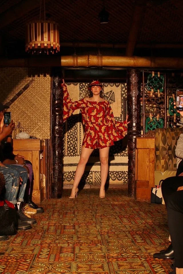 A model enters the runway wearing a short red and gold dress with long sleeves and exposed shoulders. She wears a matching visor.