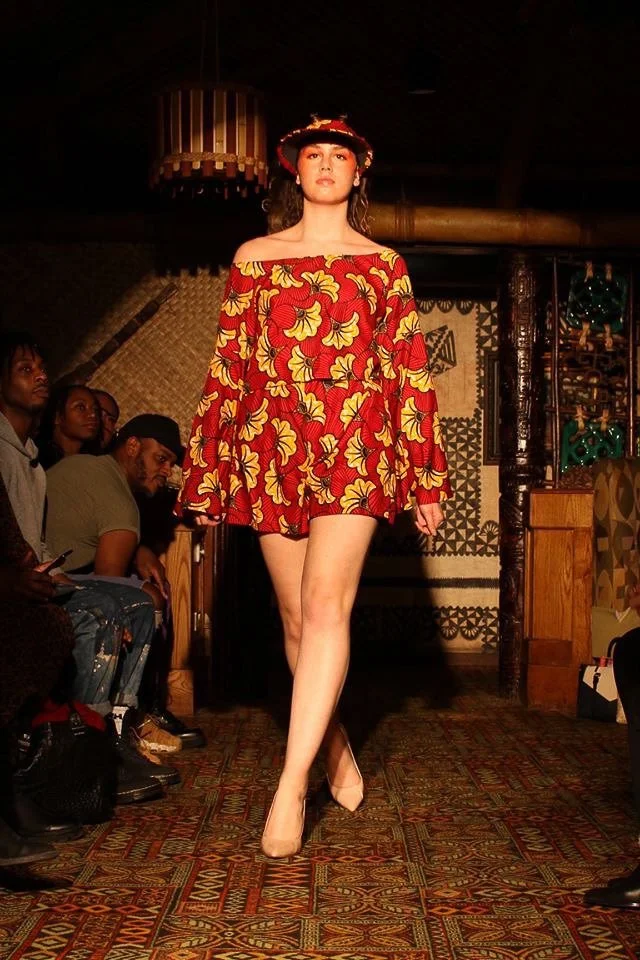 A model poses on the runway wearing a short red and gold dress with long sleeves and exposed shoulders. She wears a matching visor.