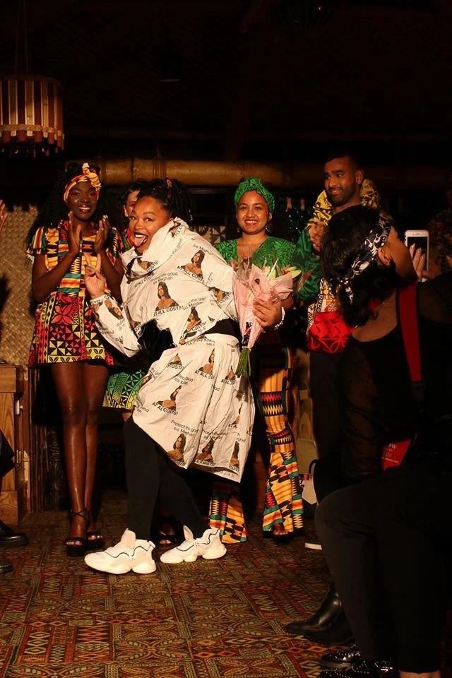 A group of models stand together in colorful garments.  Afatasi the Artist poses jubilantly in the foreground with flowers in her hand.