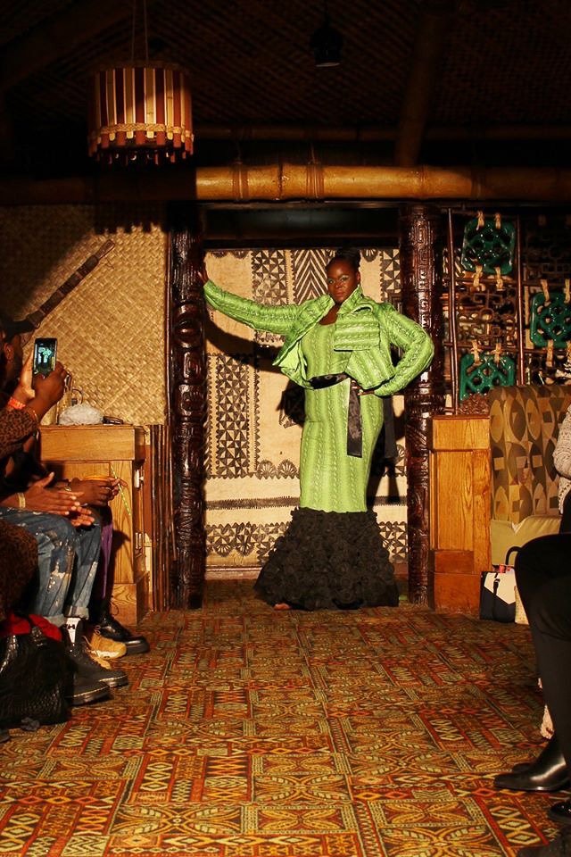 A black, female model walks the runway wearing a fitted, green mermaid dress with black ruffles.