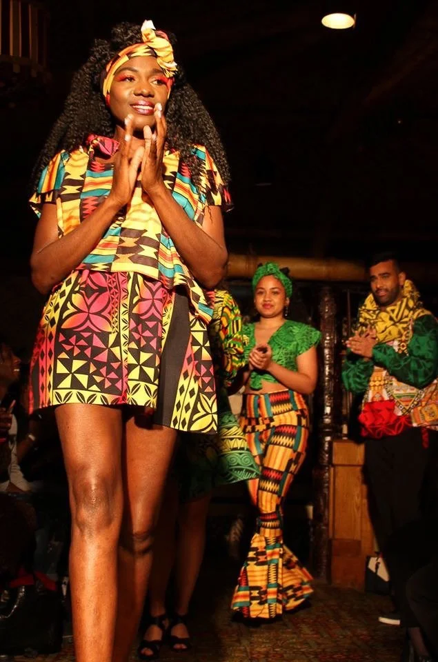Models wearing yellow, green, and red patterned garments  walk the runway together.