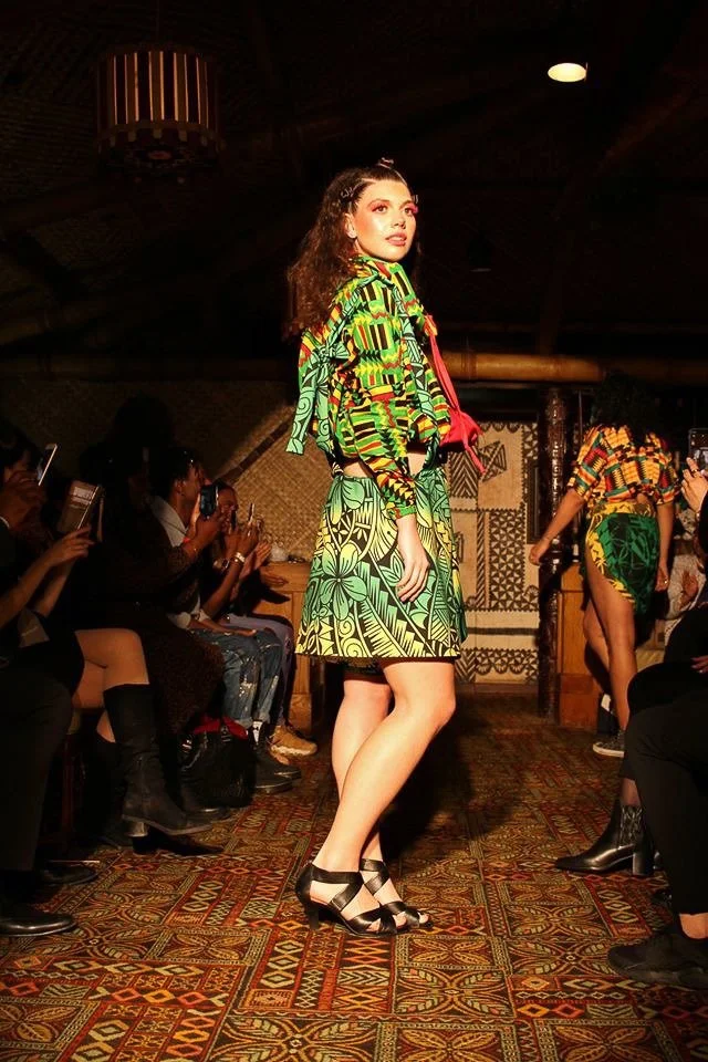 A model poses on the runway wearing a green and yellow patterned skirt with matching top. A red color frames her neck.