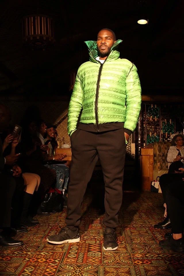 A black, male model poses on the runway wearing a puffy green coat with a hoody