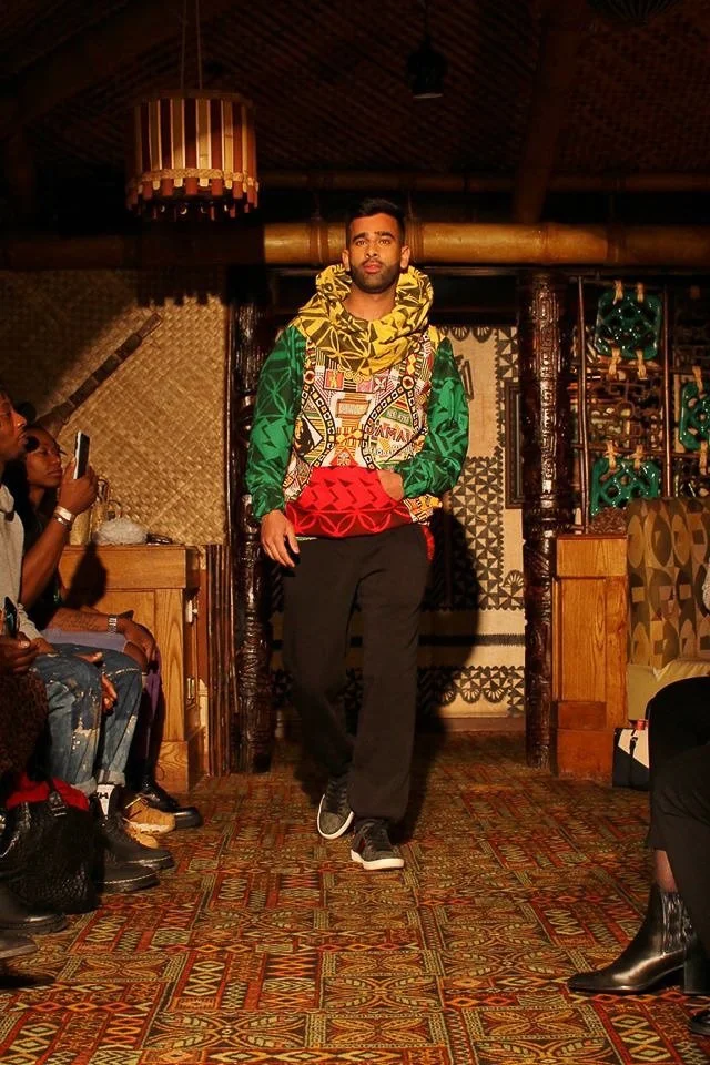 A black, male model struts on the runway showcasing a silky green, red, and yellow pullover.