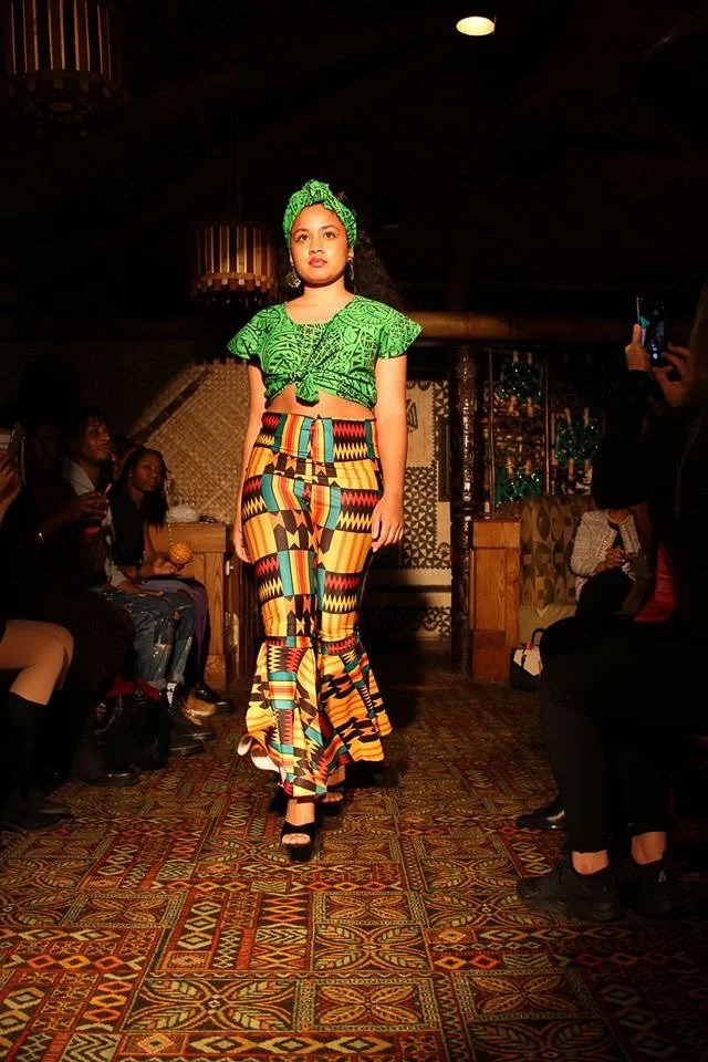 A model walks the runway wearing a green top and matching head band. She also wears fitted bell-bottom pants with green, red, gold, and black detailing.