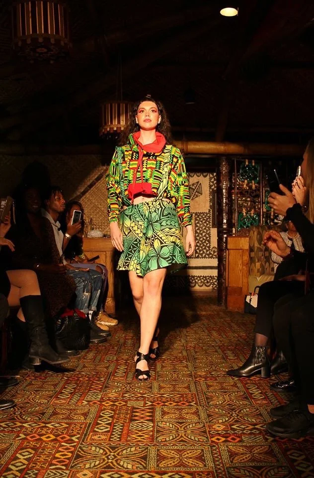 A model walks the runway wearing a green and yellow patterned skirt with matching top. A red color frames her neck.