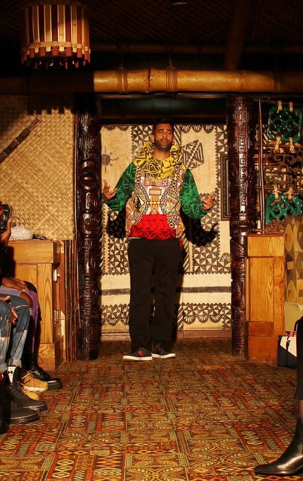 A black, male model poses on the runway showcasing a silky green, red, and yellow pullover.