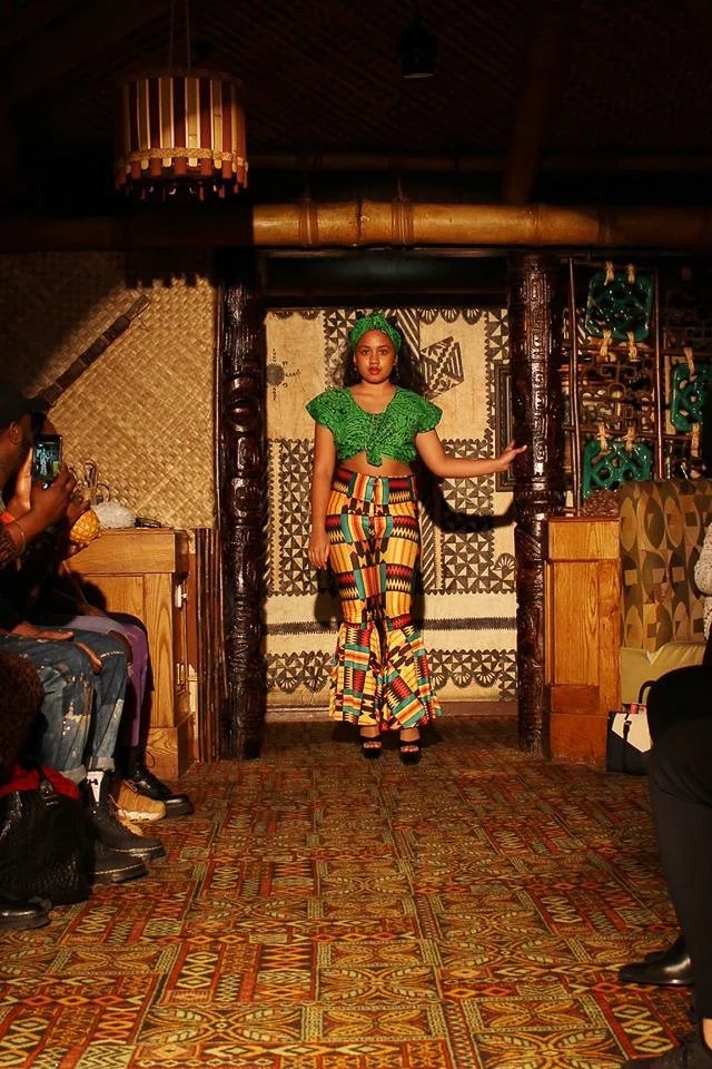 A model enters the runway wearing a green top and matching head band. She also wears fitted bell-bottom pants with green, red, gold, and black detailing.