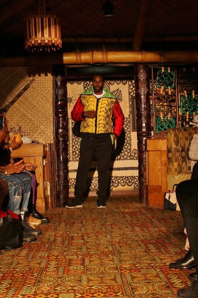 A black, male model poses on the runway showcasing a silky green, red, and yellow pullover.