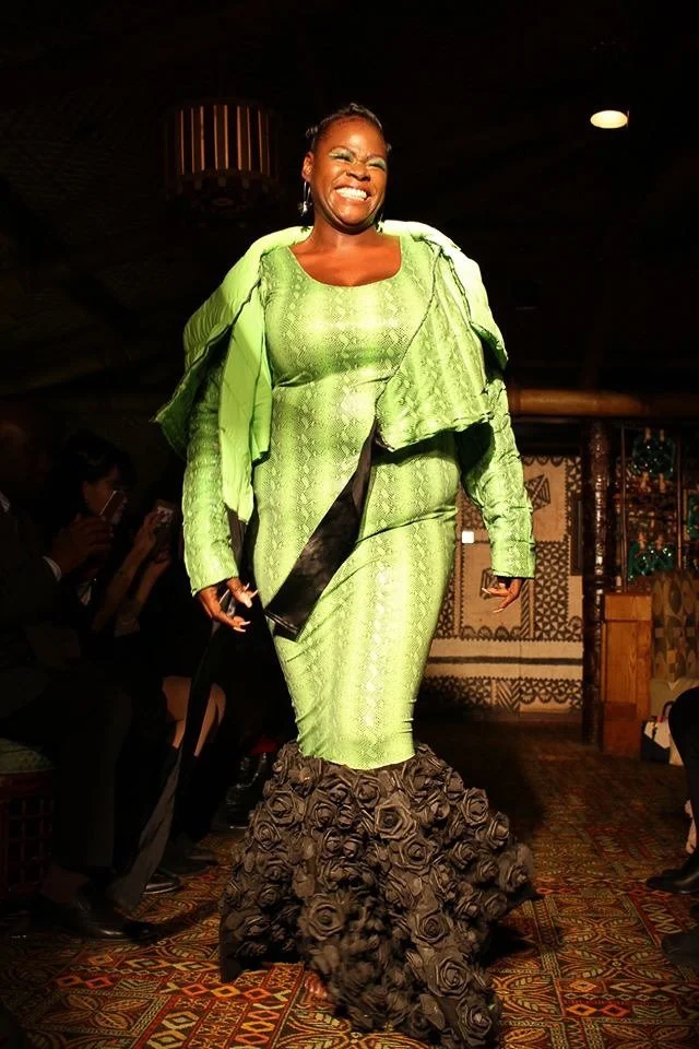 A black, female model walks the runway wearing a fitted, green mermaid dress with black ruffles.