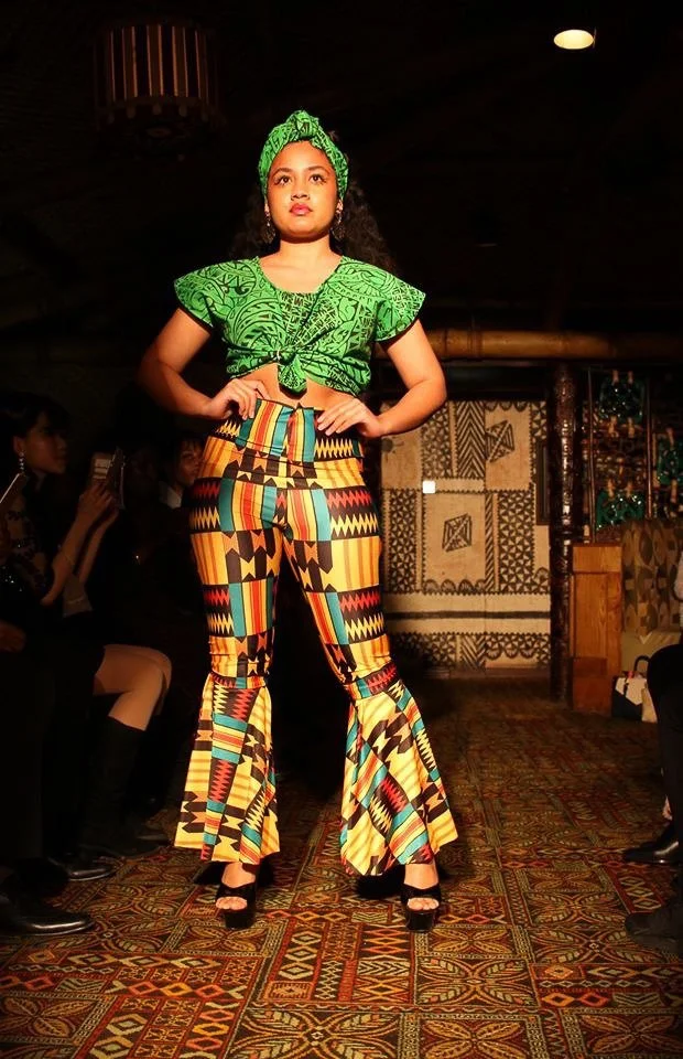 A model poses on the runway wearing a green top and matching head band. She also wears fitted bell-bottom pants with green, red, gold, and black detailing.