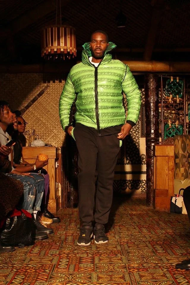 A black, male model walks the runway wearing a puffy green coat with a hoody