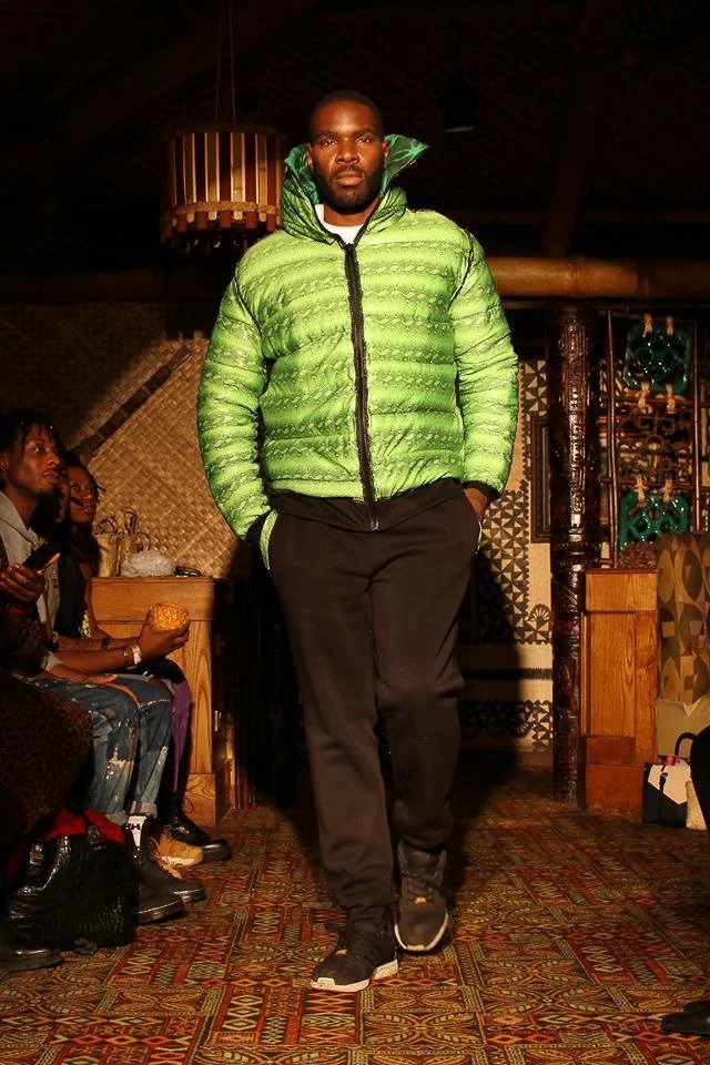 A black, male model walks the runway wearing a puffy green coat with a hoody