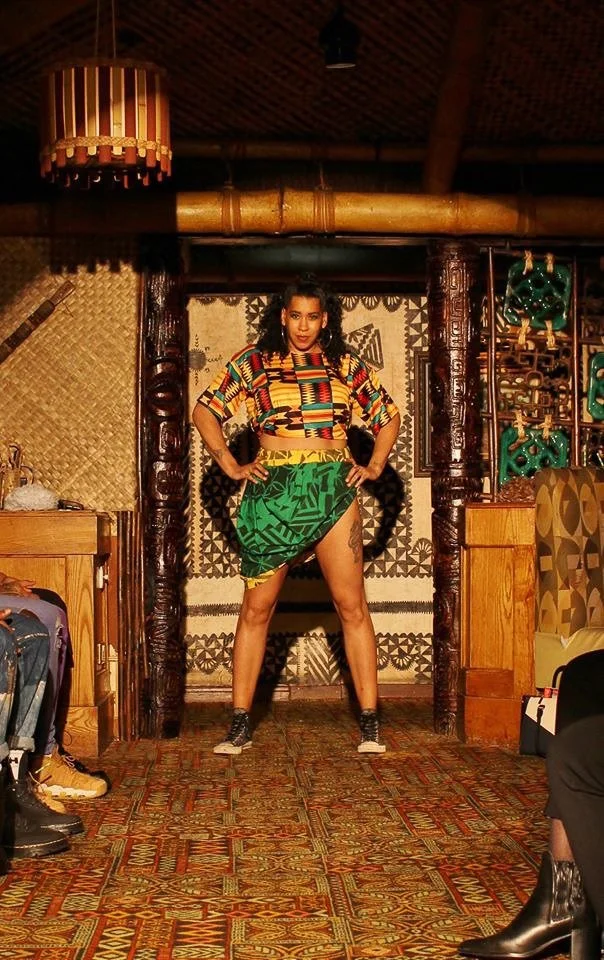 A black, female model strikes a pose wearing a geometric patterned yellow, red, and green top over a shiny green and yellow, asymmetrical skirt. 