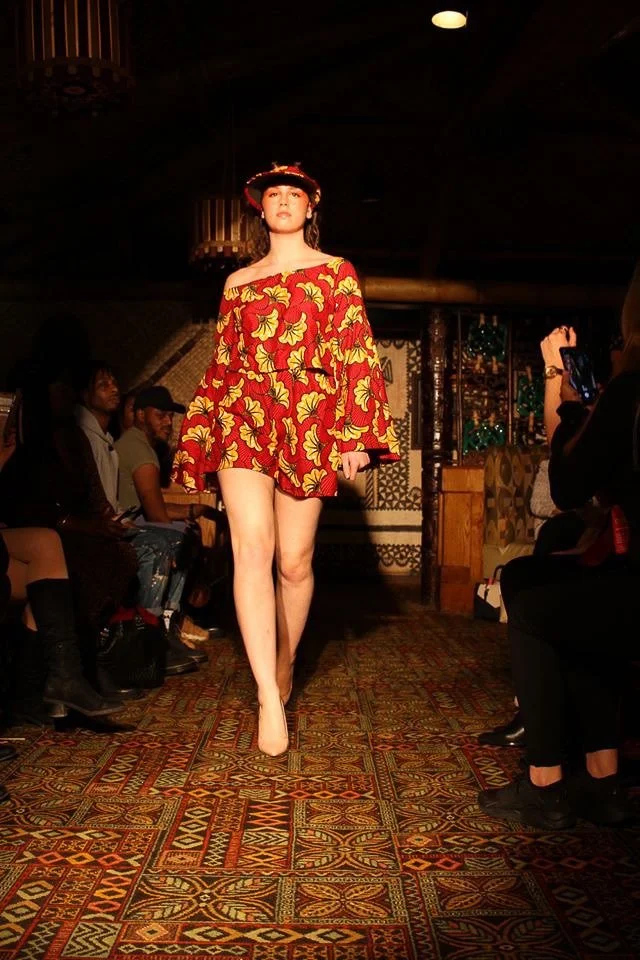 A model walks the runway wearing a short red and gold dress with long sleeves and exposed shoulders. She wears a matching visor.
