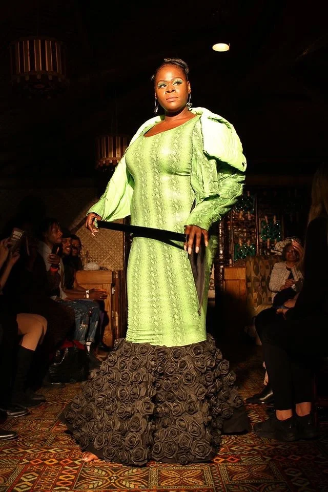 A black, female model walks the runway wearing a fitted, green mermaid dress with black ruffles.
