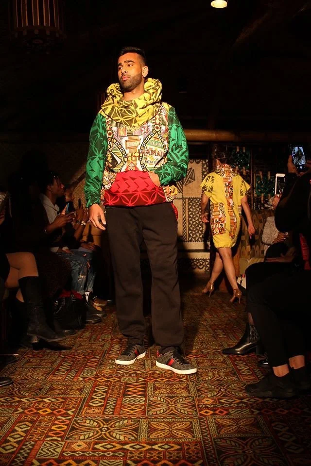 A black, male model poses on the runway showcasing a silky green, red, and yellow pullover.