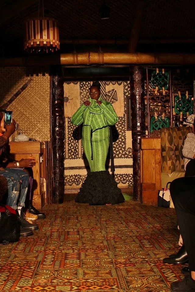 A black, female model walks the runway wearing a fitted, green mermaid dress with black ruffles.