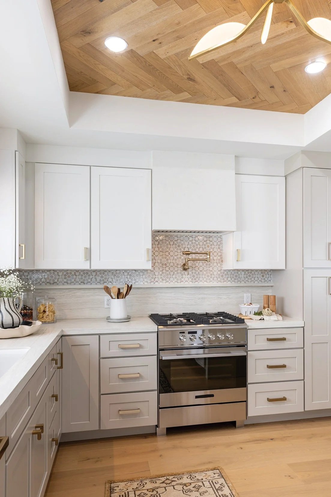 A modern kitchen with white cabinets, a stainless steel stove, and wood flooring. The ceiling features a recessed wooden design with built-in lights and a gold-colored chandelier.