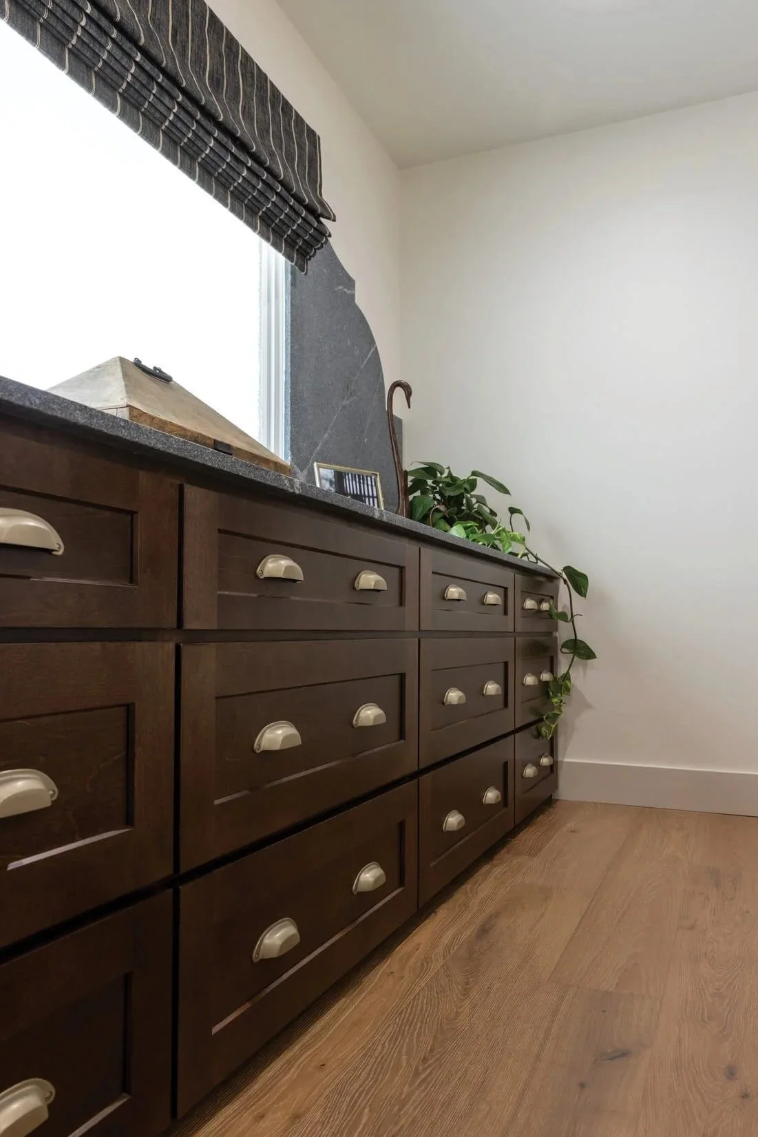 A dark brown wooden dresser with multiple drawers, cream-colored handles, situated against a white wall; atop the dresser are a small decorative box, a framed photo, and a potted plant, near a window with a dark striped valance.