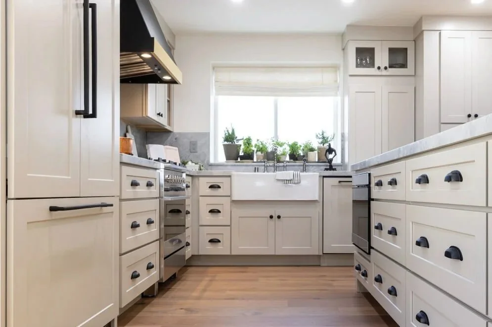 Bright white kitchen with wooden floors, multiple drawers and cabinets with black handles, a kitchen island, a window behind the sink with potted plants, and a vase with orange flowers on the counter.