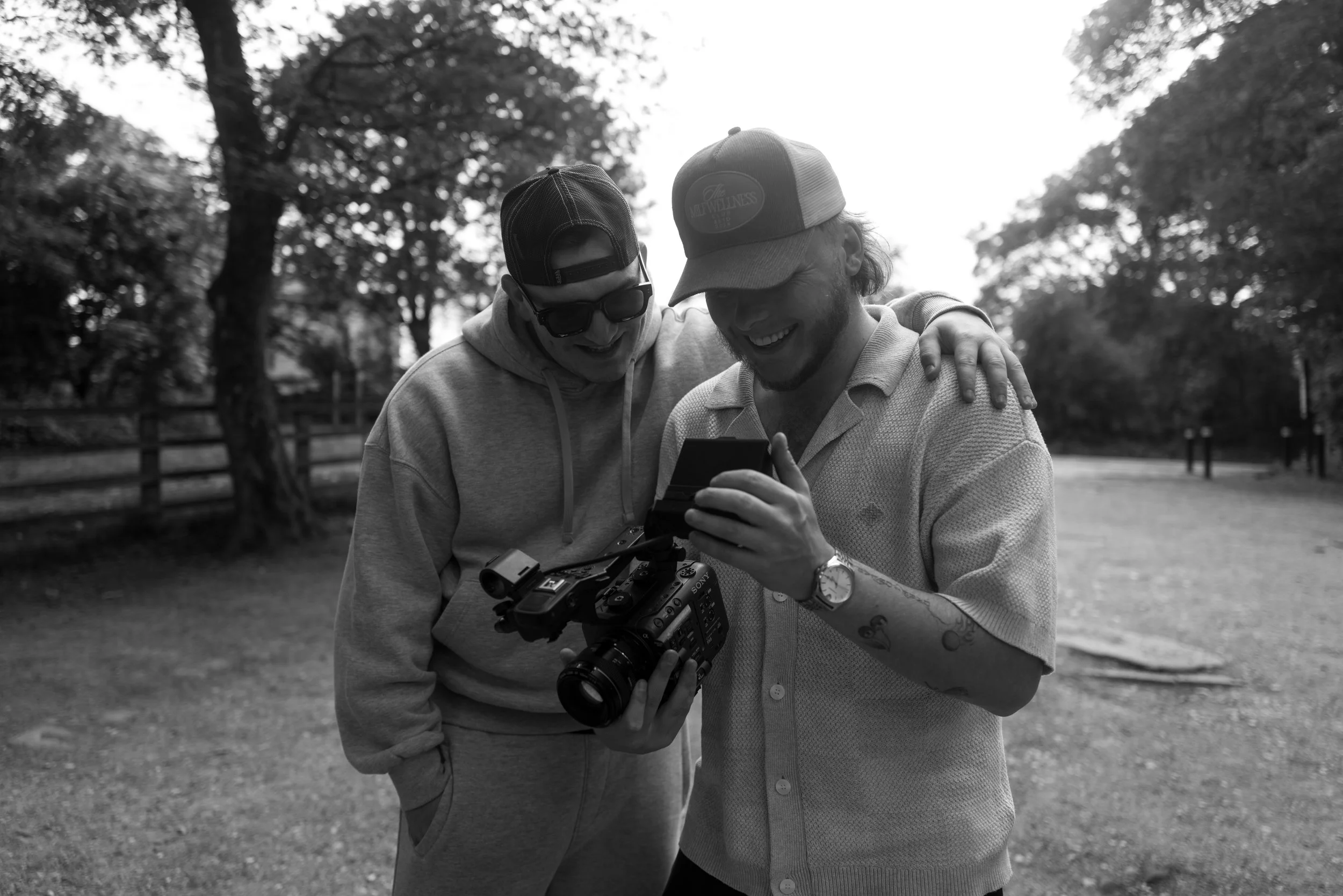 Two young men laughing and looking at a camera and phone outdoors in a park.