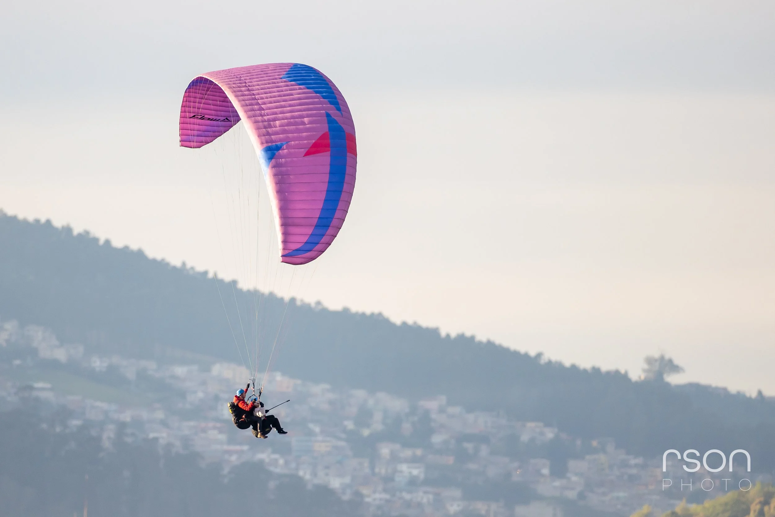 A person paragliding with a pink and purple canopy over a landscape with hills and buildings in the background.