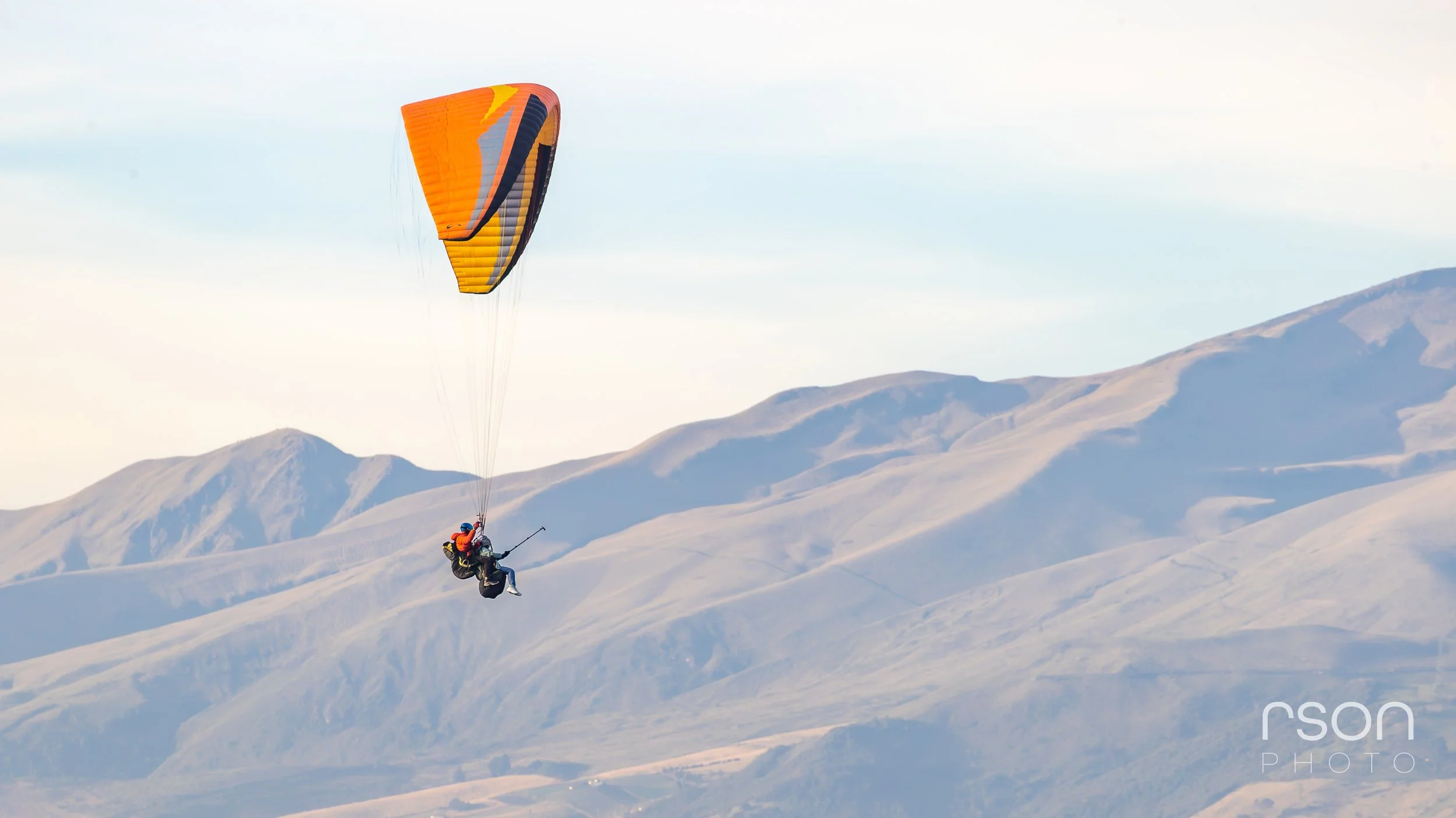 A person paragliding over a mountain range with hills and a blue sky in the background.