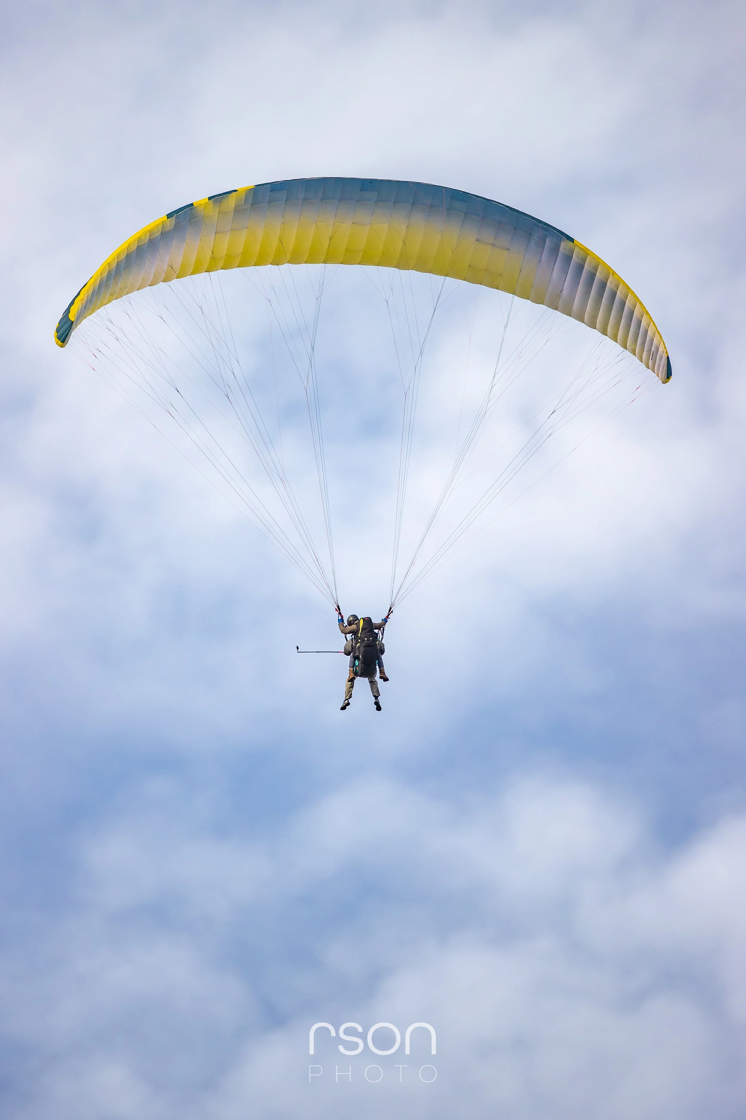 Two paragliders in mid-air with yellow and gray parachute against a cloudy sky.