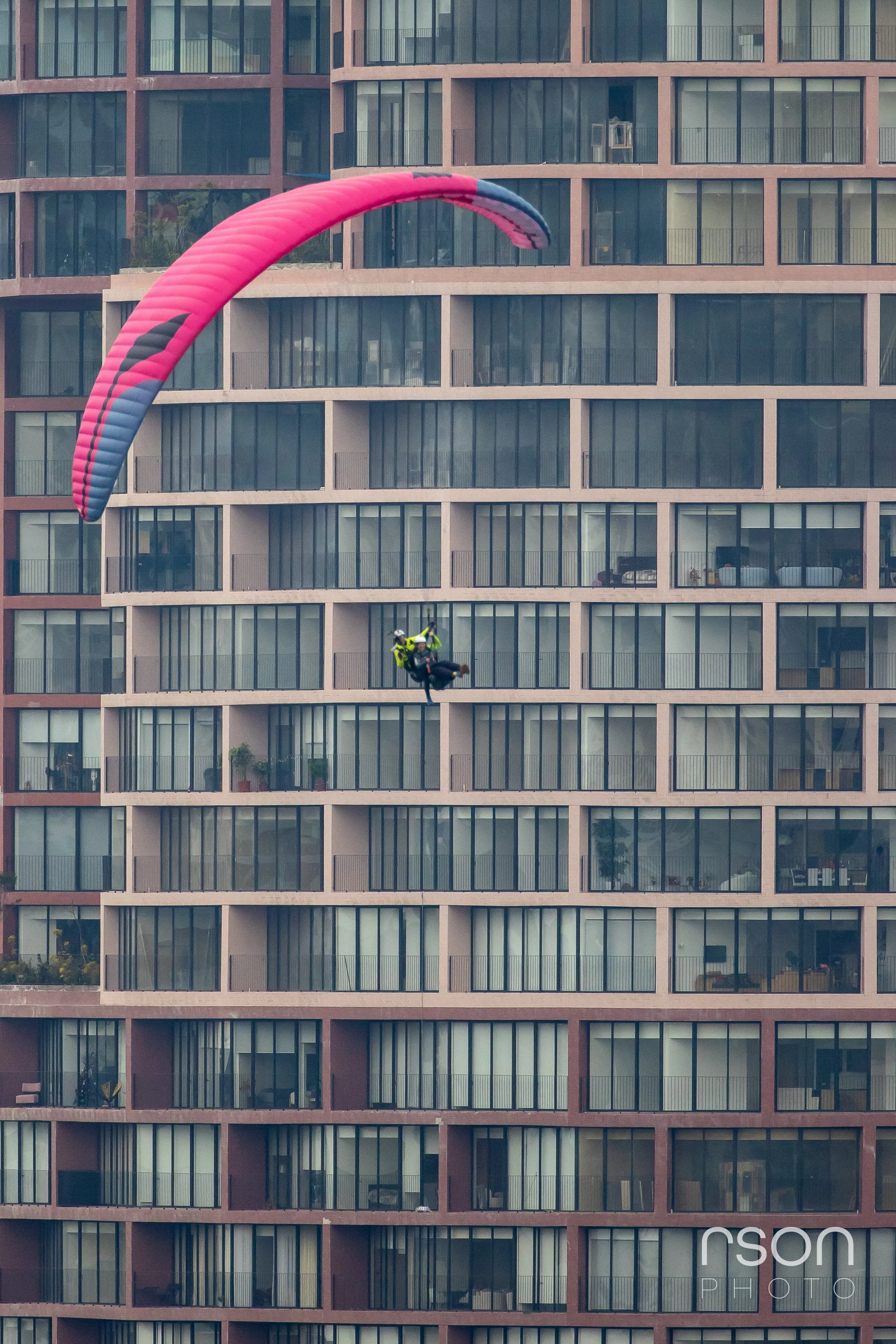A person paragliding with a pink and blue canopy in front of a tall apartment building.