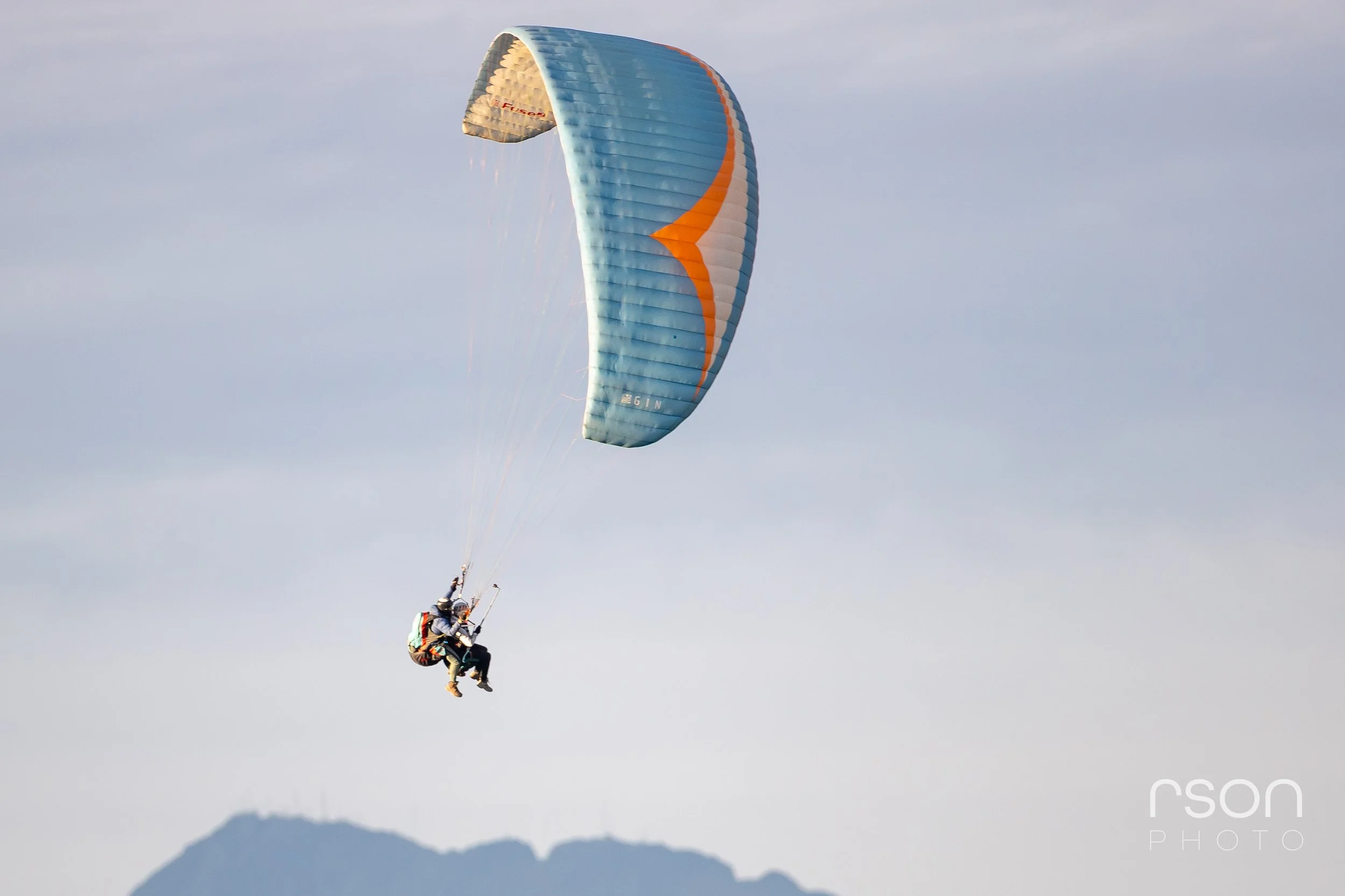Paraglider in mid-air against cloudy sky with mountain silhouette below.