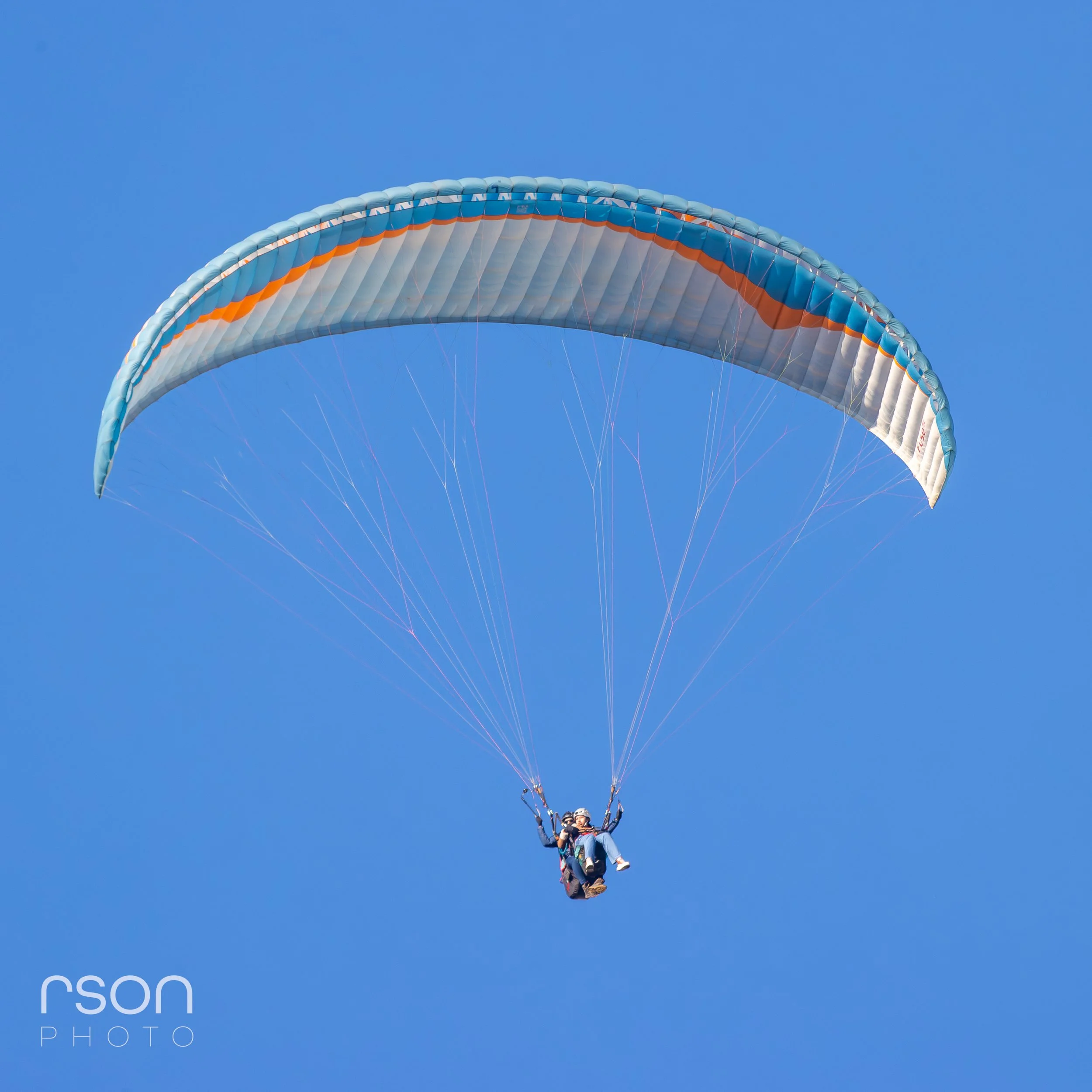 A person paragliding in clear blue sky with a silver and orange parachute.