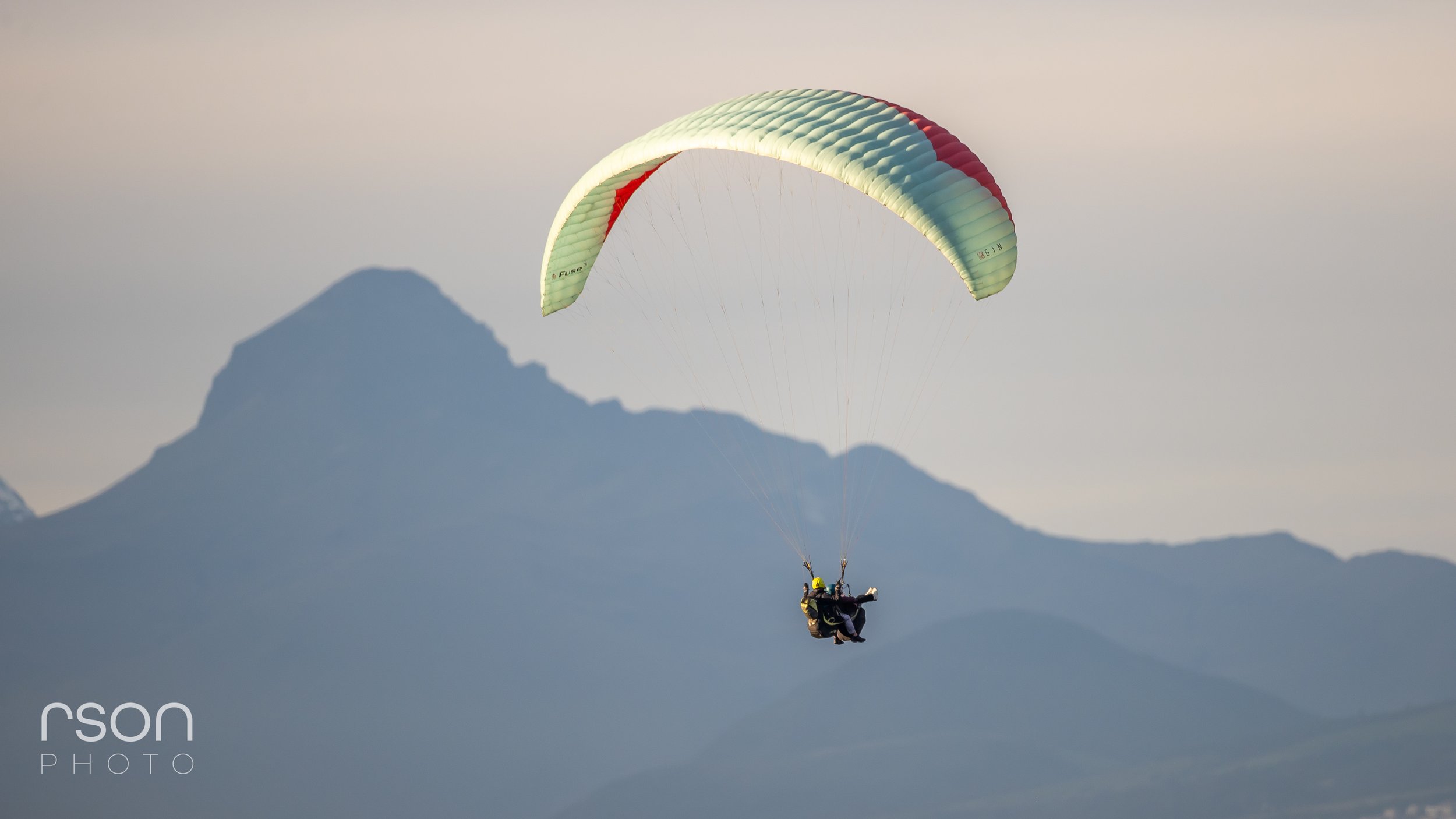 A person paragliding with a light green parachute against a mountain backdrop and a cloudy sky.