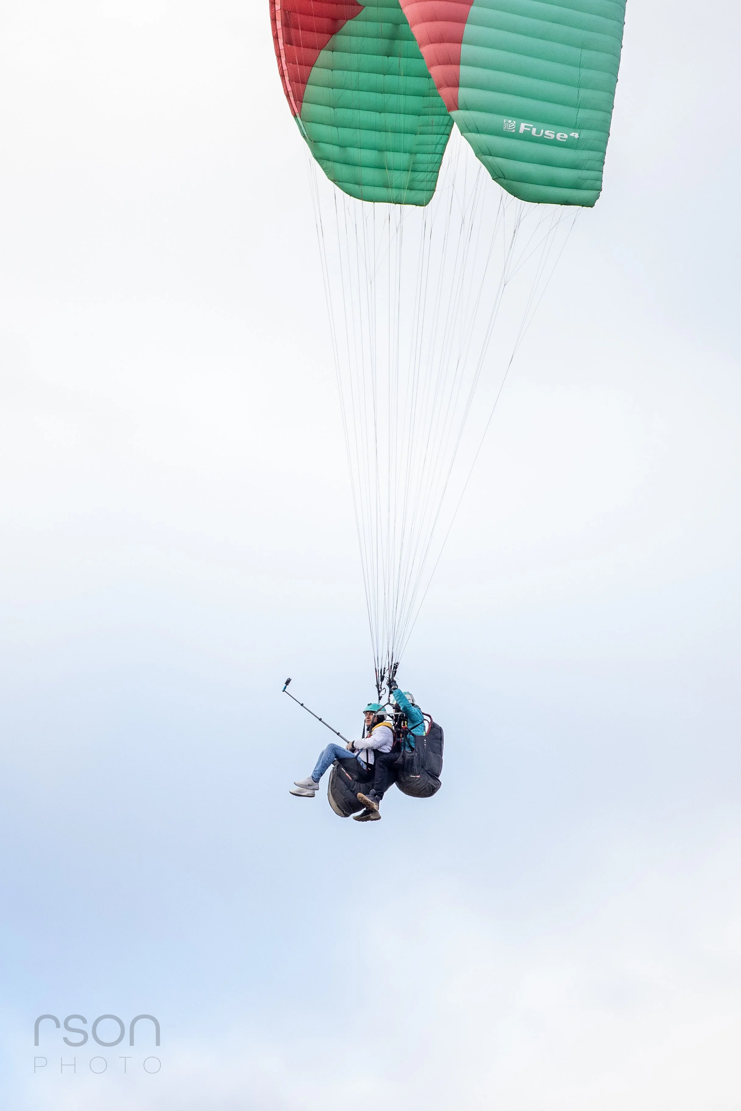 Two people in a tandem paraglider flying through the sky with a green and red parachute canopy.
