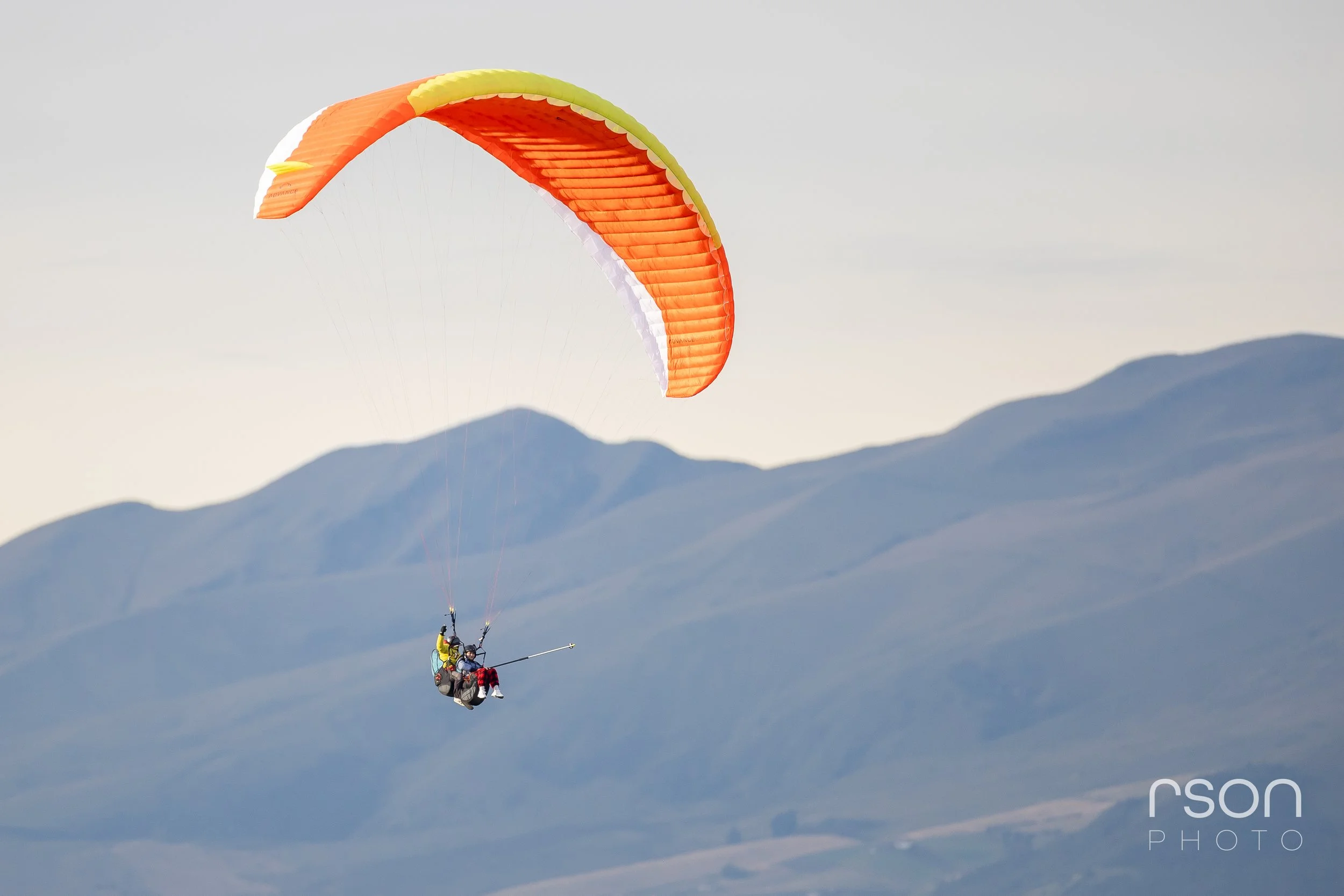A person in a harness flying a colorful orange and yellow paraglider in the sky over mountains.