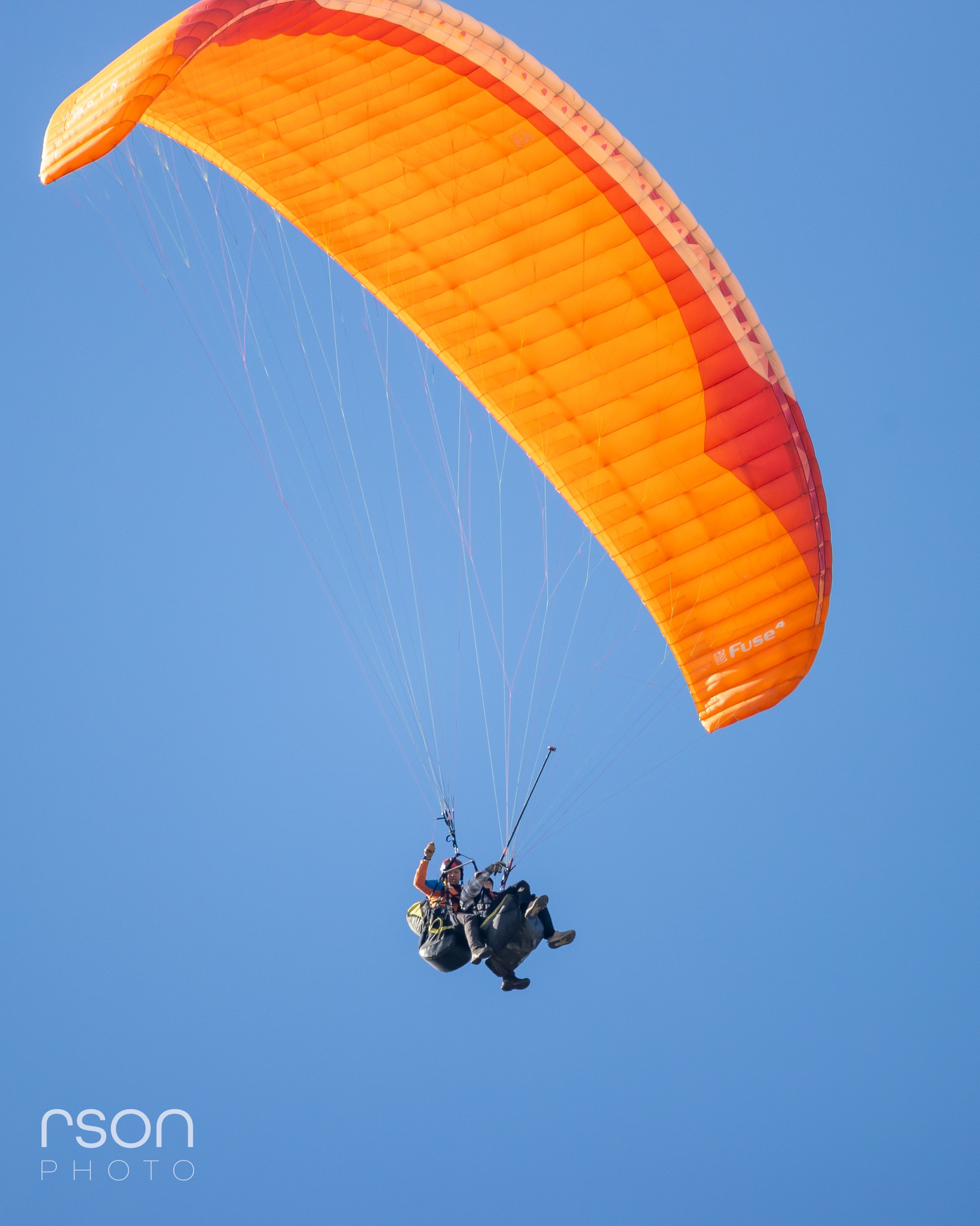 A person paragliding with an orange canopy against a clear blue sky.