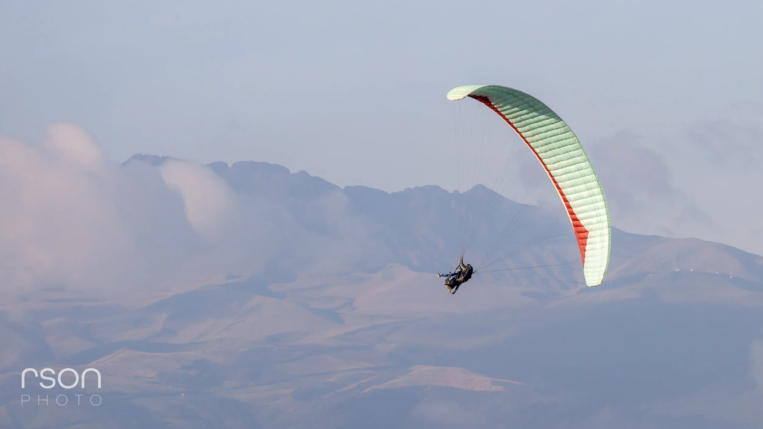 A person paragliding in the sky above mountains and clouds, with a green and red parachute.