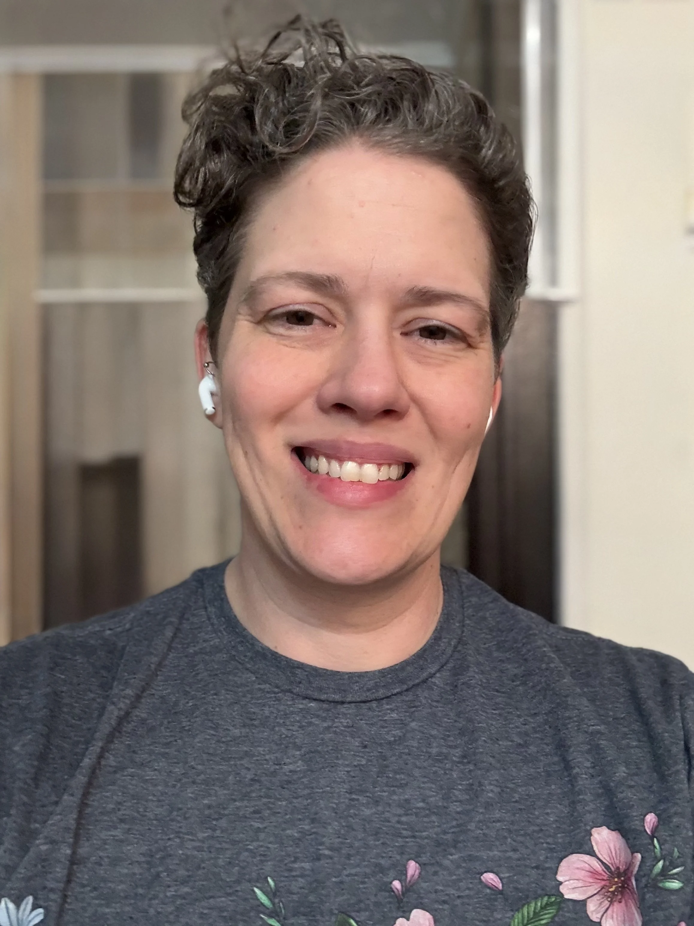 A person smiling, wearing a gray T-shirt with pink floral designs, wireless earbuds, and short curly hair, indoors.
