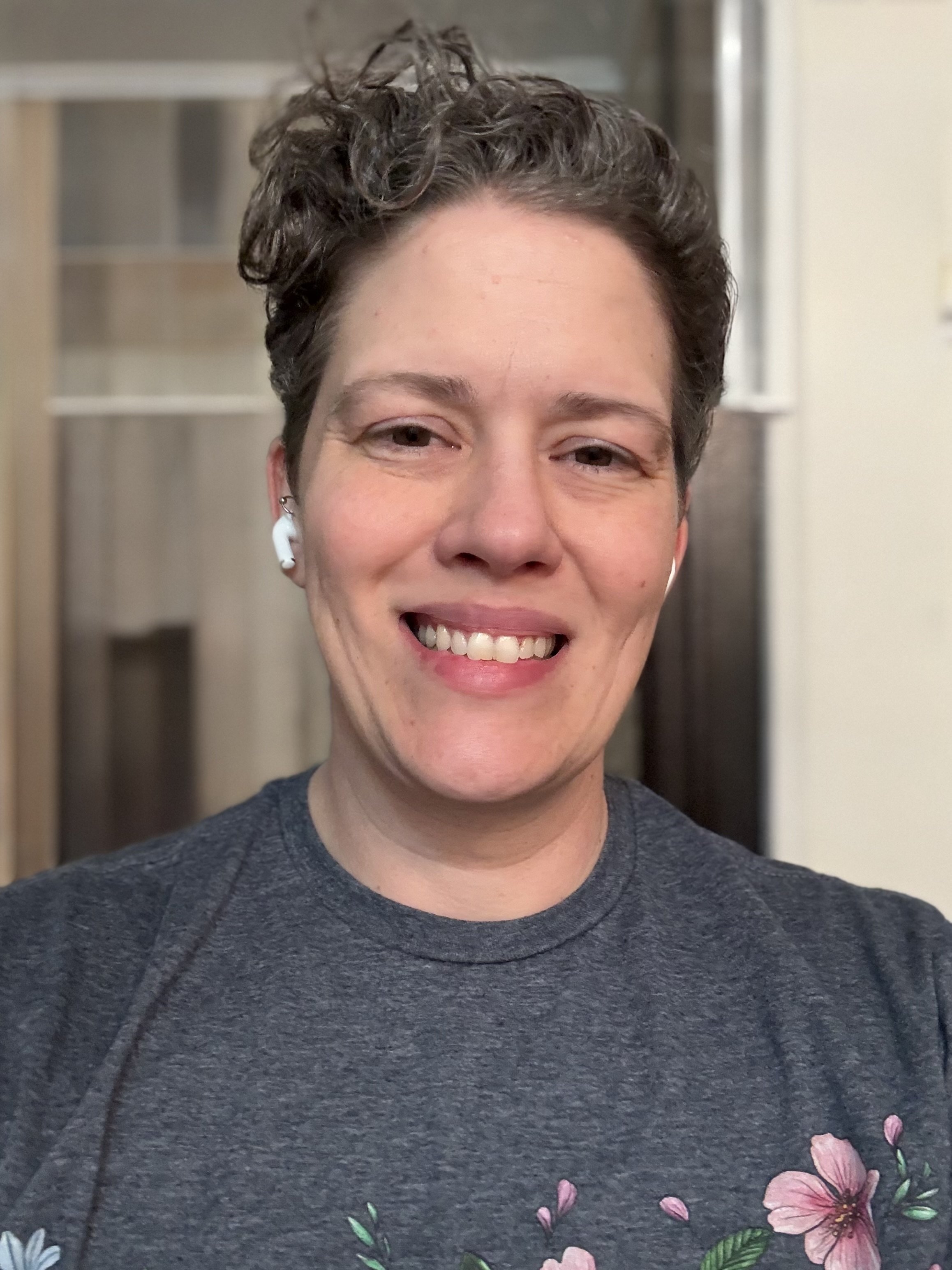 A person smiling, wearing a gray T-shirt with pink floral designs, wireless earbuds, and short curly hair, indoors.