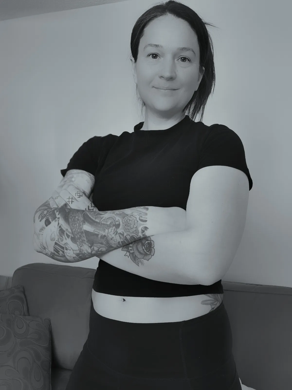 A woman with short dark hair, wearing a black T-shirt and dark pants, standing with arms crossed in a room with plain light-colored walls and a patterned sofa in the background. She has tattoos on her arms and a belly button piercing.