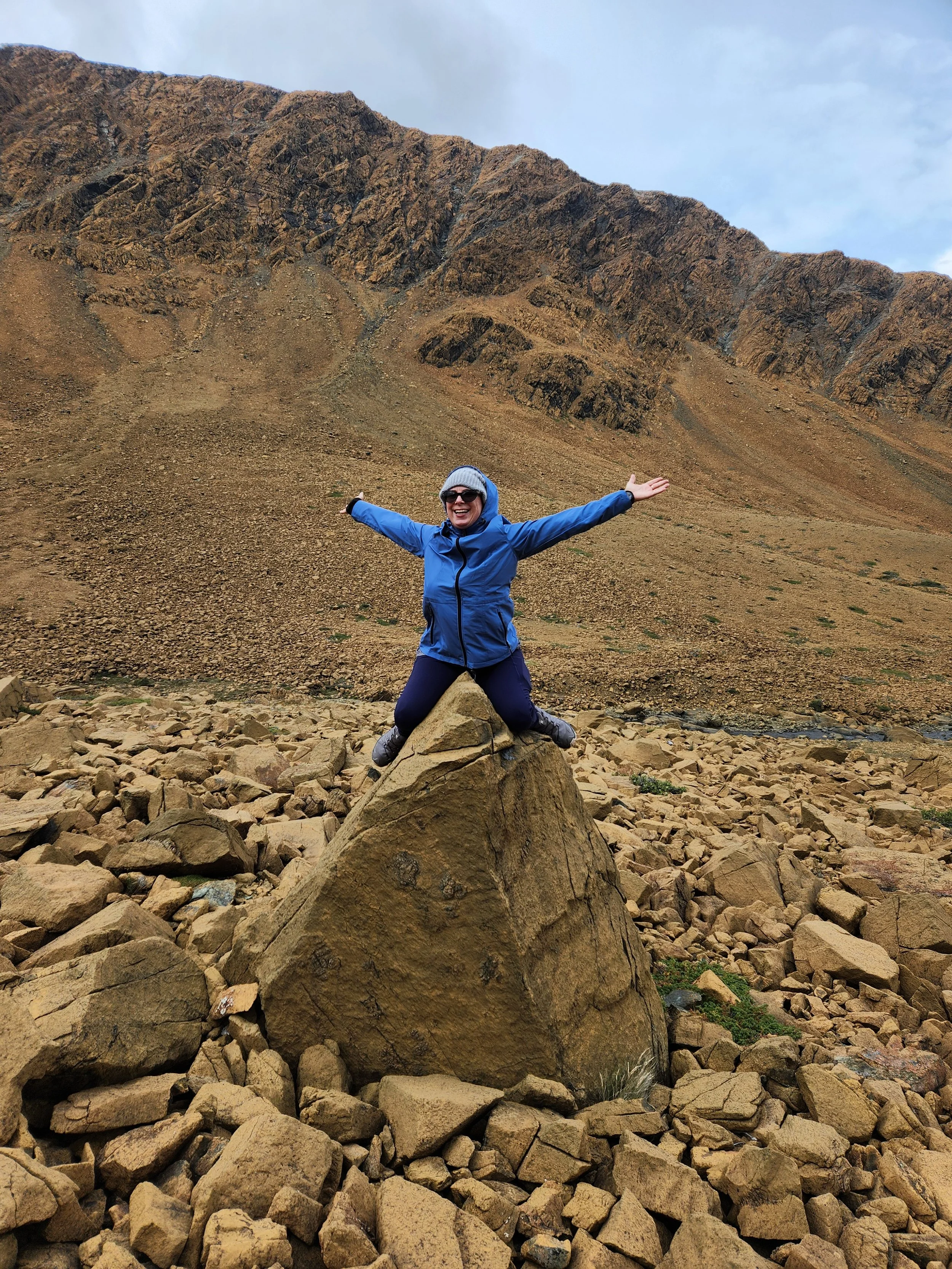 A woman wearing a blue jacket, gray hat, and sunglasses smiling and posing with arms outstretched while sitting on a large rock among rocky terrain in a mountainous area.