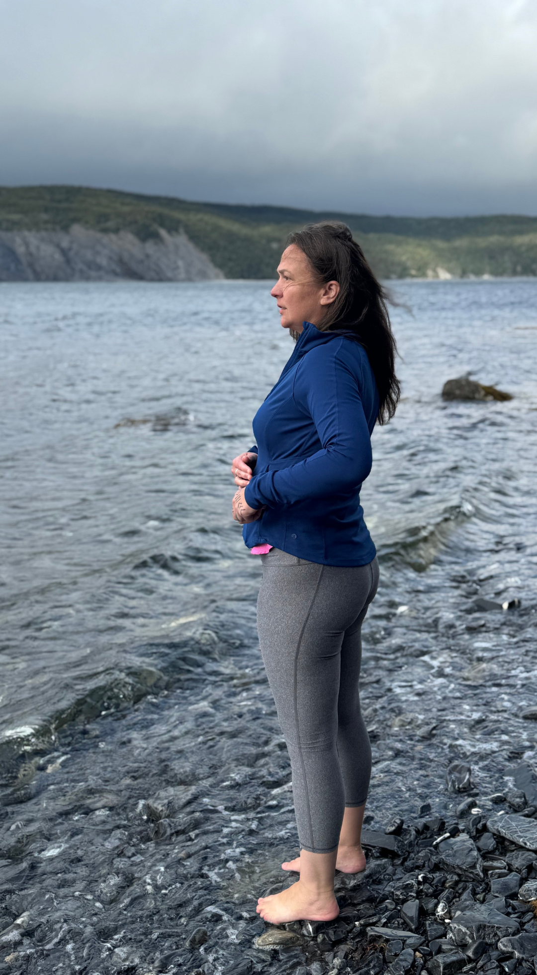 A woman standing barefoot on a rocky shoreline by a body of water, wearing a blue jacket and gray leggings, with dark hair and looking pensively into the distance.