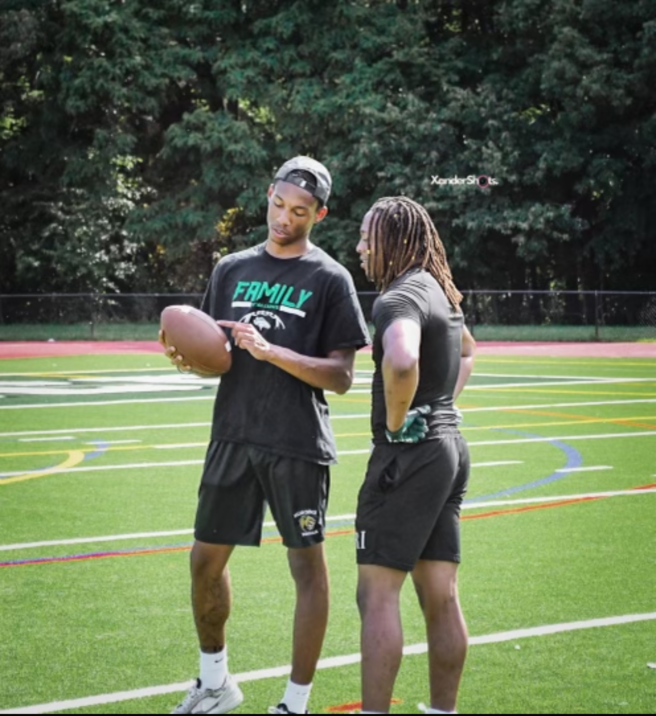 Two young men standing on a football field, one holding a football and showing something on it to the other, with trees in the background.