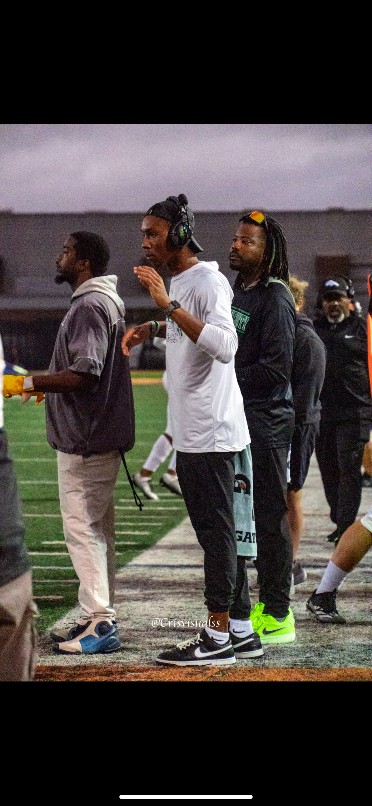 Three young men standing on a sports field, two wearing headphones, one with colorful dreadlocks and sunglasses, dressed in athletic clothing, during a game or practice session.