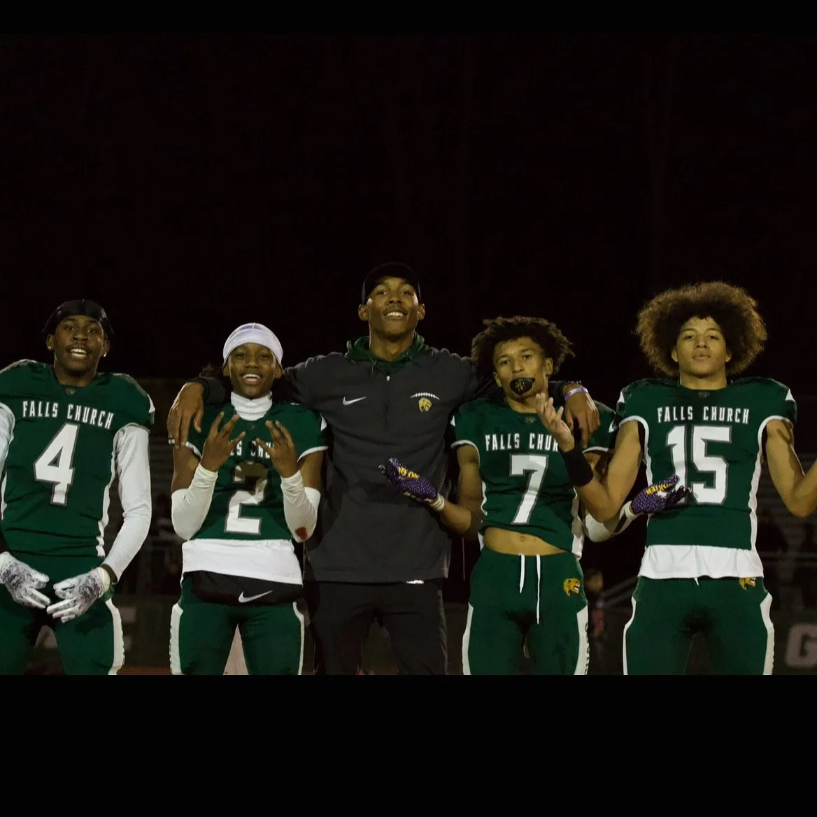 A group of five young football players in green and white uniforms with 'Falls Church' written on them, standing with a coach on a football field at night, celebrating after a game.