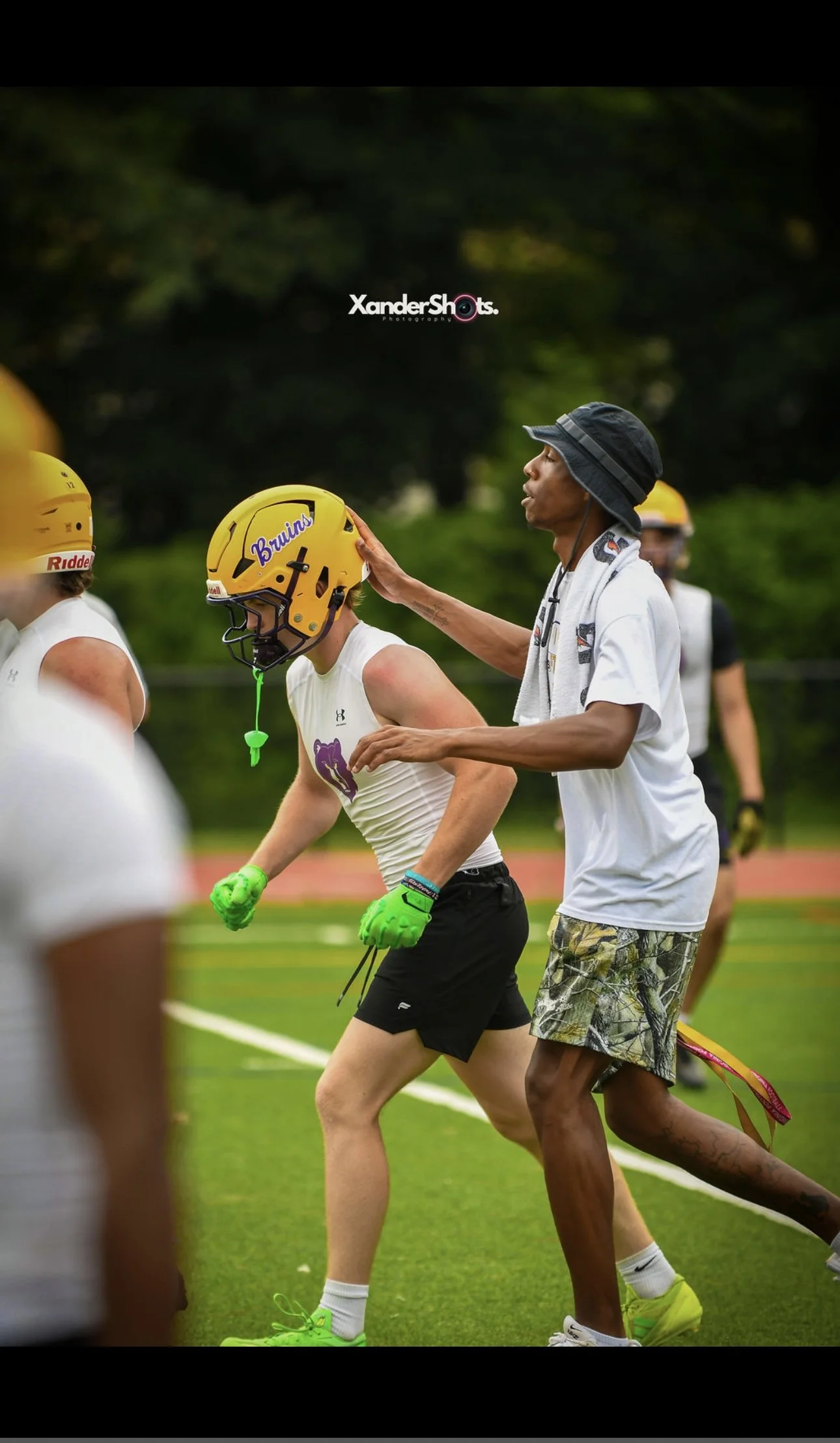 A football coach or trainer touches the helmet of a player during a practice or game, while other players in helmets and athletic gear are in the background on the field.