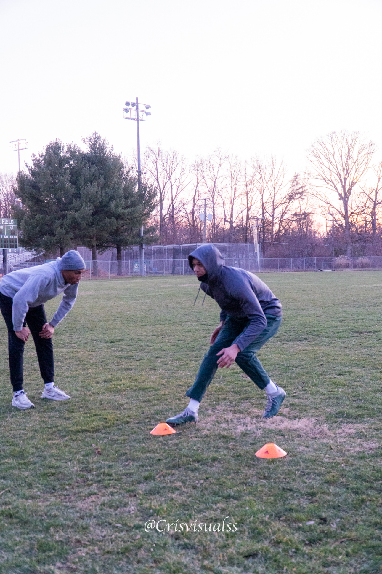 Two young men in athletic clothing and hoodies practicing on a grassy field with orange cones during sunset or early evening