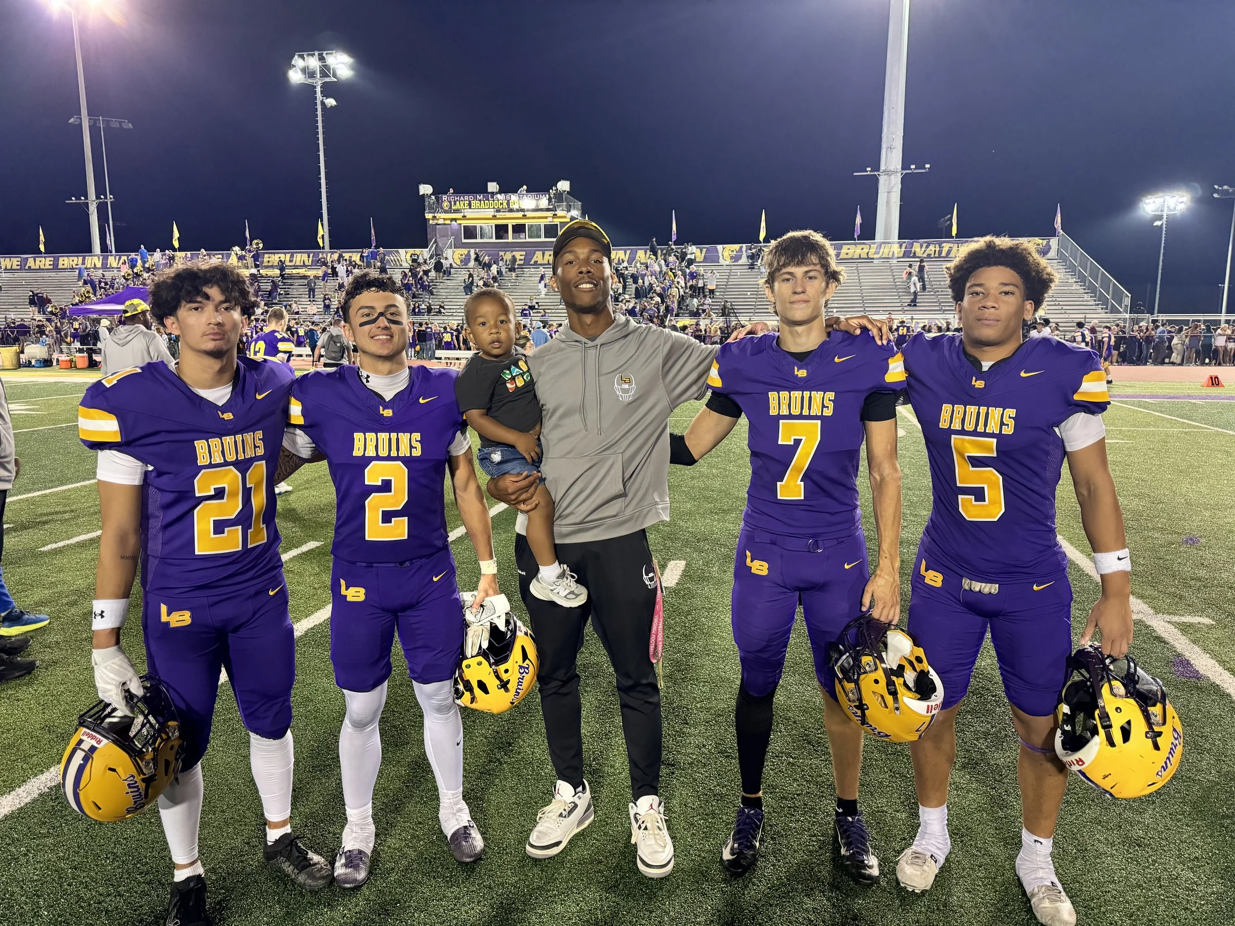 Group of five football players in purple uniforms and a coach with a baby, standing on a football field at night.