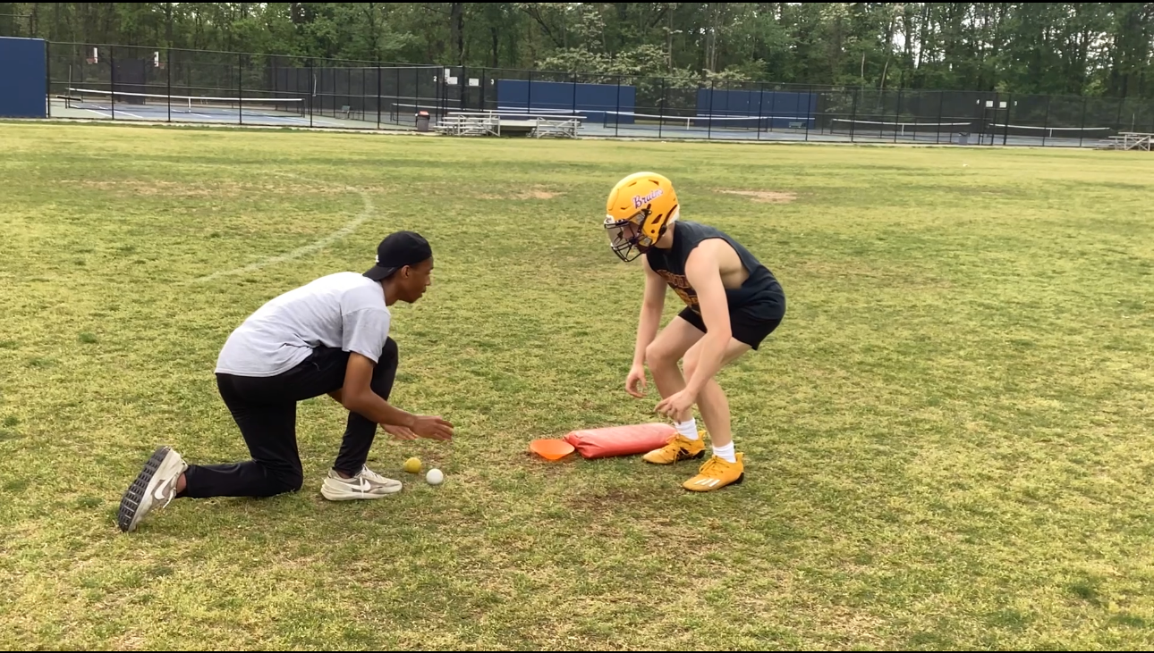 A coach and a young football player practicing on a field, with the player wearing a yellow helmet and tackling pads, and the coach kneeling next to a white tennis ball and a yellow tennis ball.